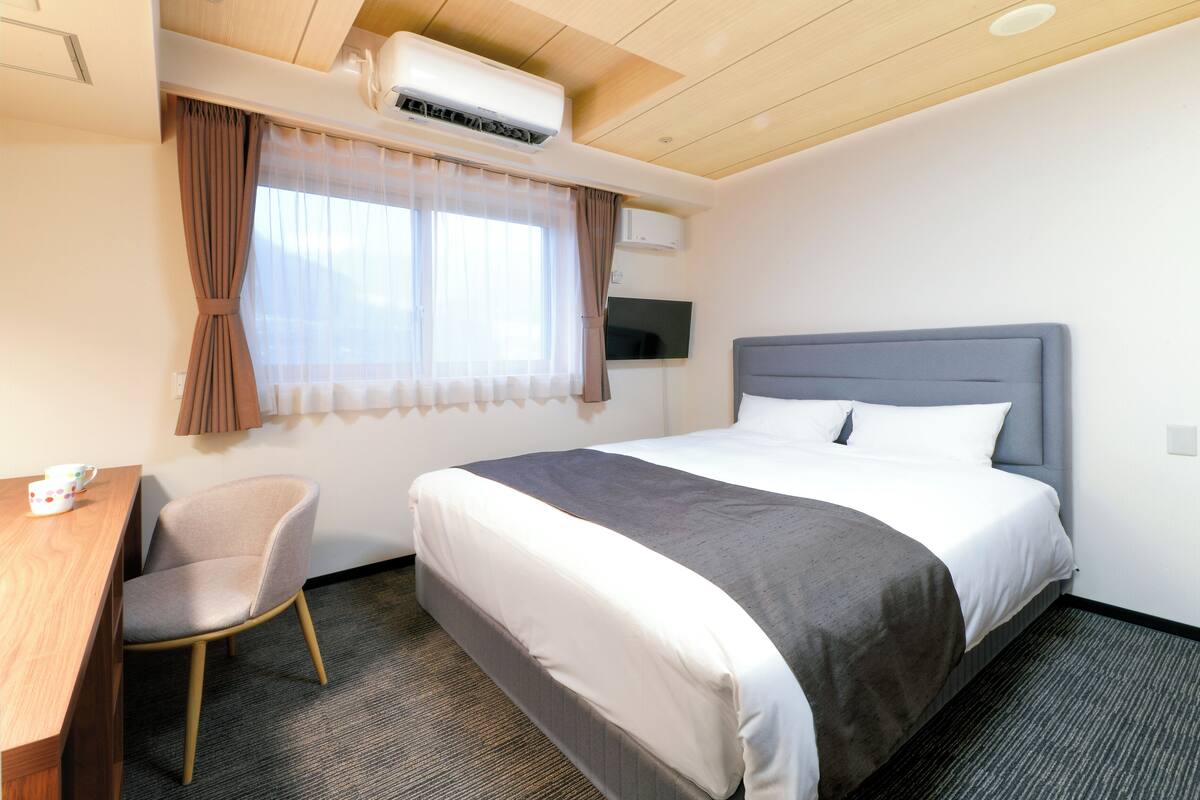 A hotel room with a bed, a desk, a chair, an air conditioning unit, and a television on the wall.