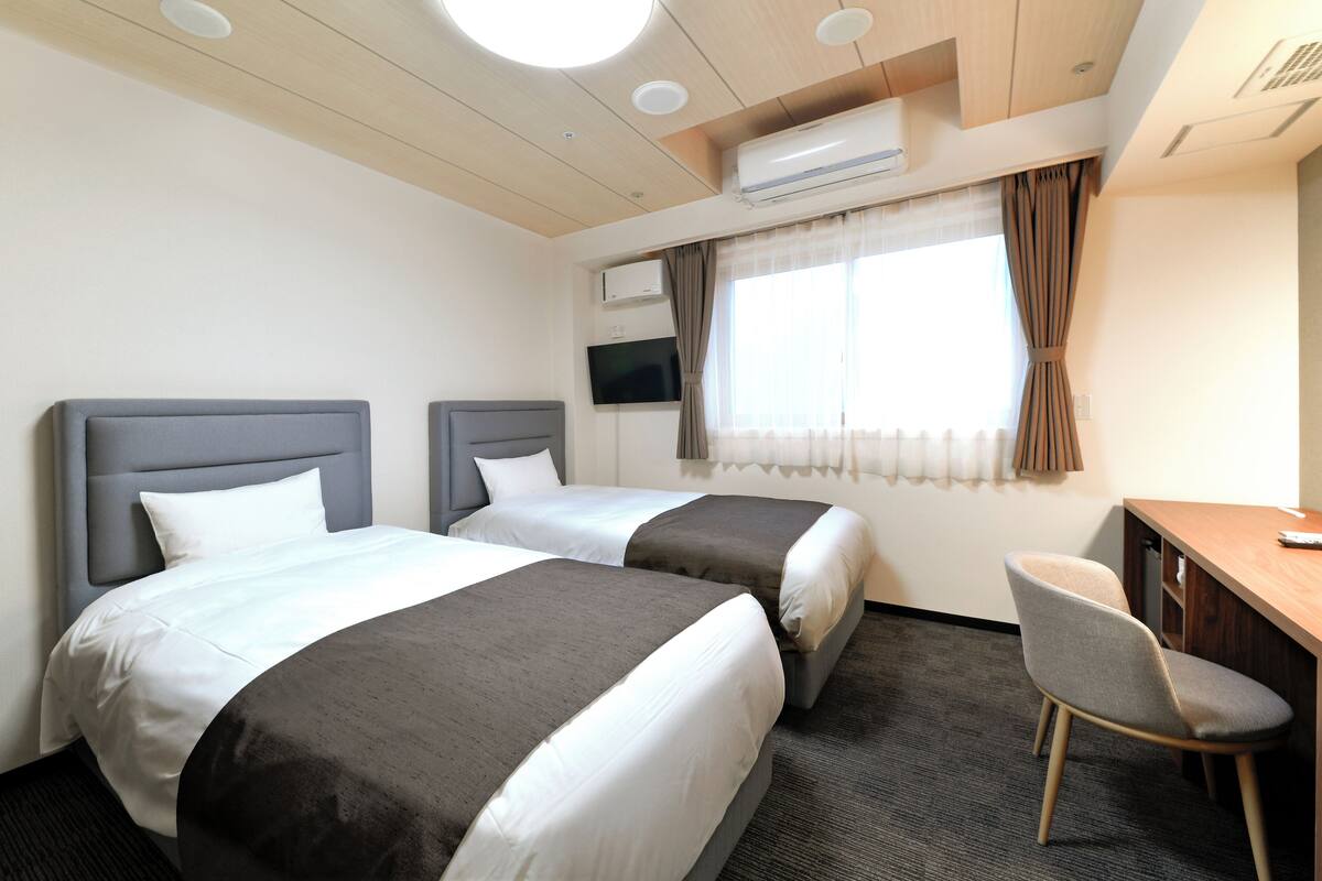 A hotel room with two beds, a desk, a chair, a TV, and an air conditioning unit.