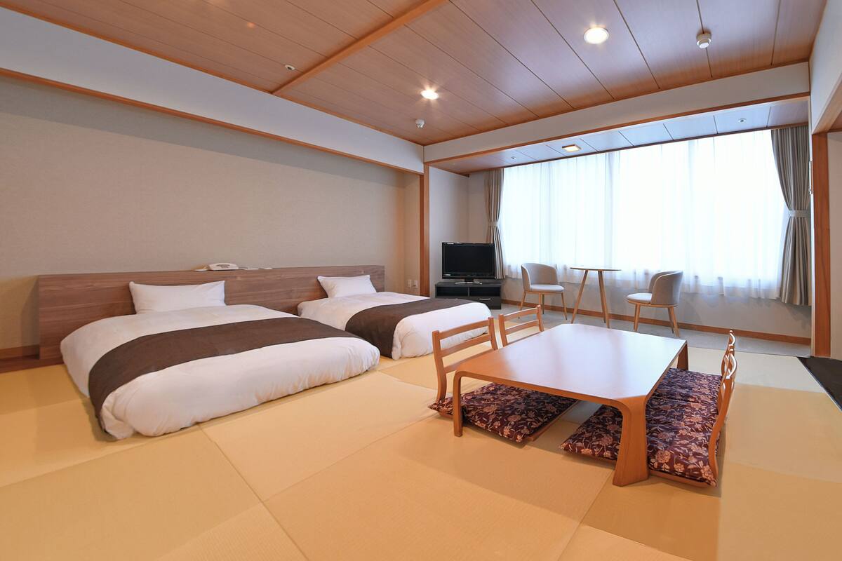 A hotel room with two beds, a TV, a dining table, and a view of the outside.