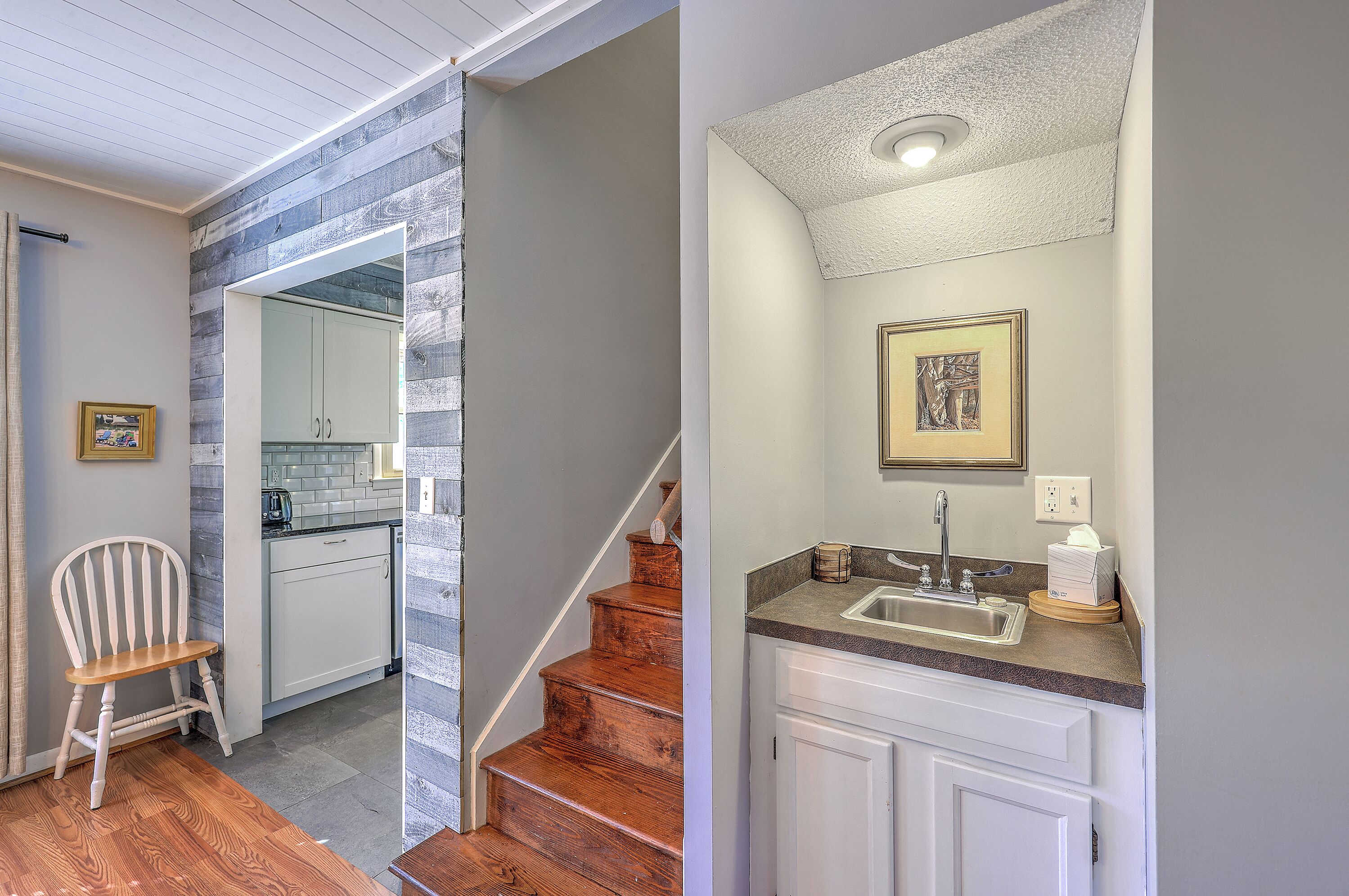 A modern kitchen with a wooden staircase, a dining area with a wooden chair, and a bathroom with a sink and towel dispenser.