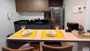 A compact kitchen with a refrigerator, stove, and sink. There are two dining tables with yellow placemats and white plates. - Solon Sales Luxor Prime (João Pessoa)