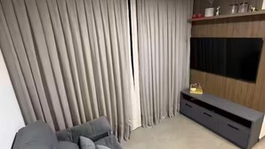 A modern living room with a grey sofa, a flat-screen TV mounted on the wall, and a large window with grey curtains. - Solon Sales Luxor Prime (João Pessoa)