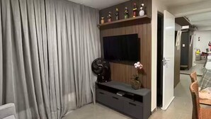 A modern living room with a flat-screen TV, a dark entertainment unit, a fan, and a dining area with a table and chairs. - Solon Sales Luxor Prime (João Pessoa)