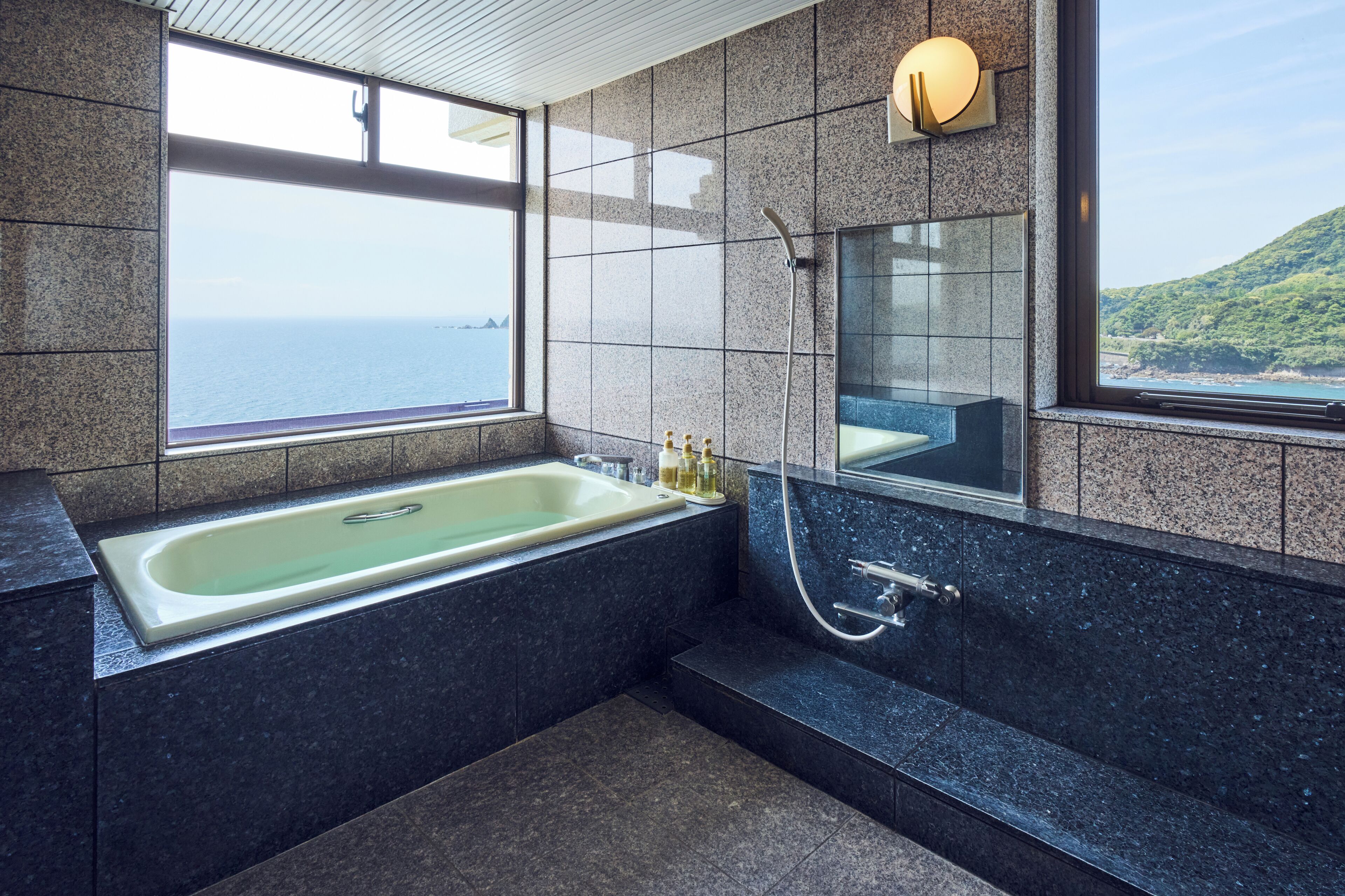 A modern bathroom with a large bathtub, a shower, and a large mirror. There are two windows providing a view of the sea and a hill.