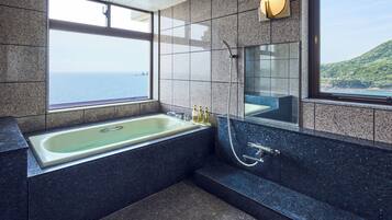 A modern bathroom with a large bathtub, a shower, and a large mirror. There are two windows providing a view of the sea and a hill.