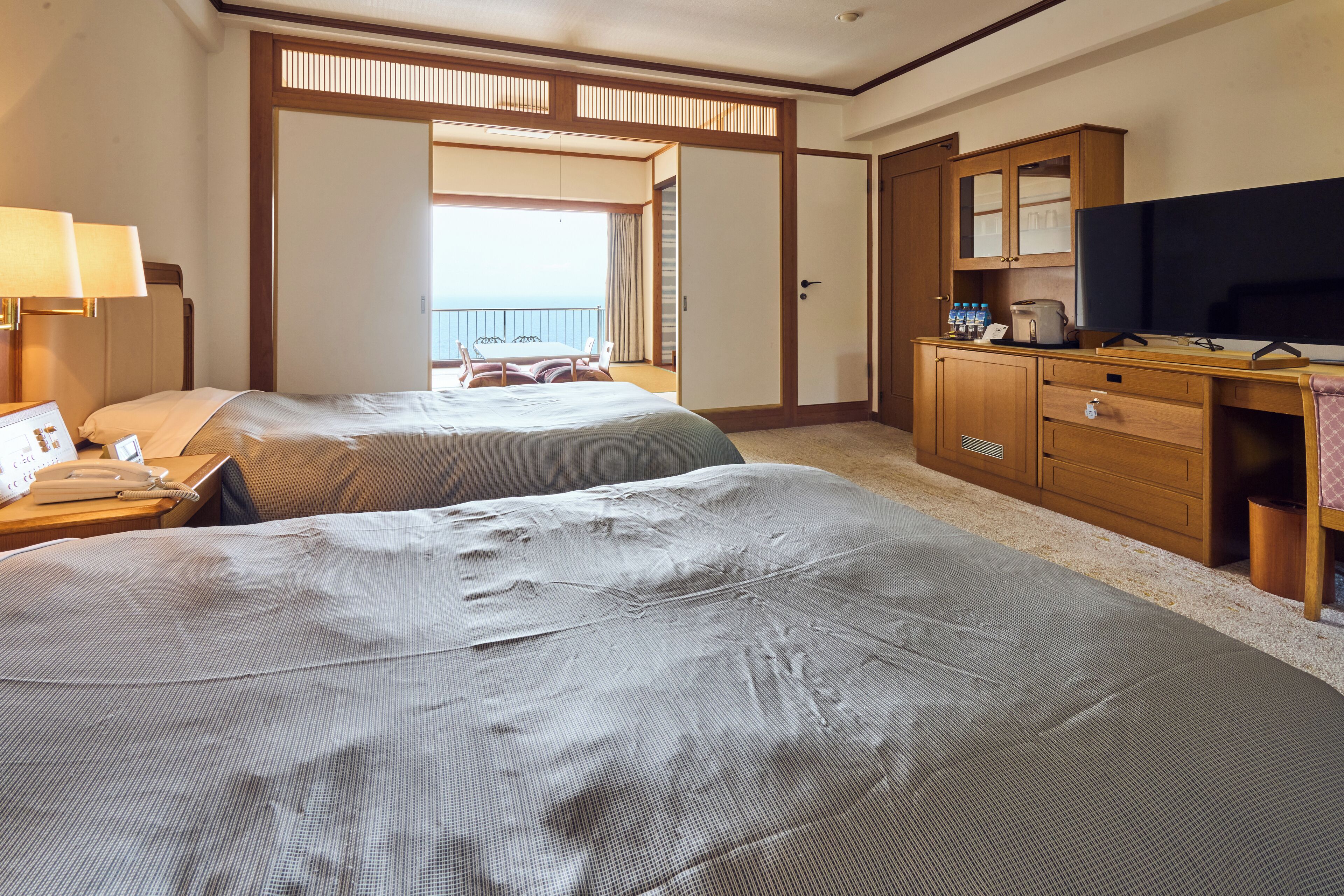 A hotel room with two beds, a desk with a TV, a minibar, and a view of the balcony.
