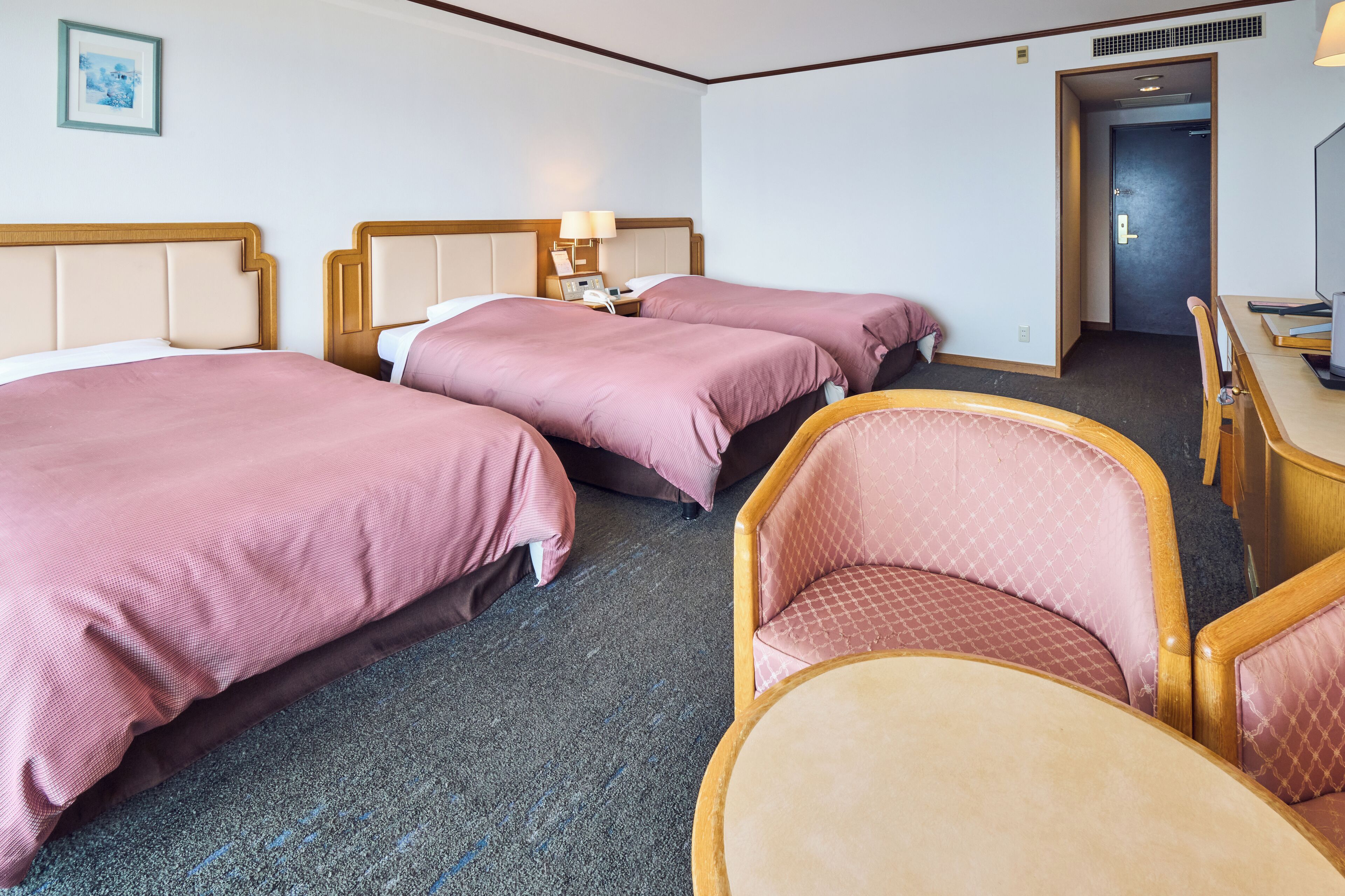 A hotel room with three beds, a desk, a chair, and a door leading to another room.