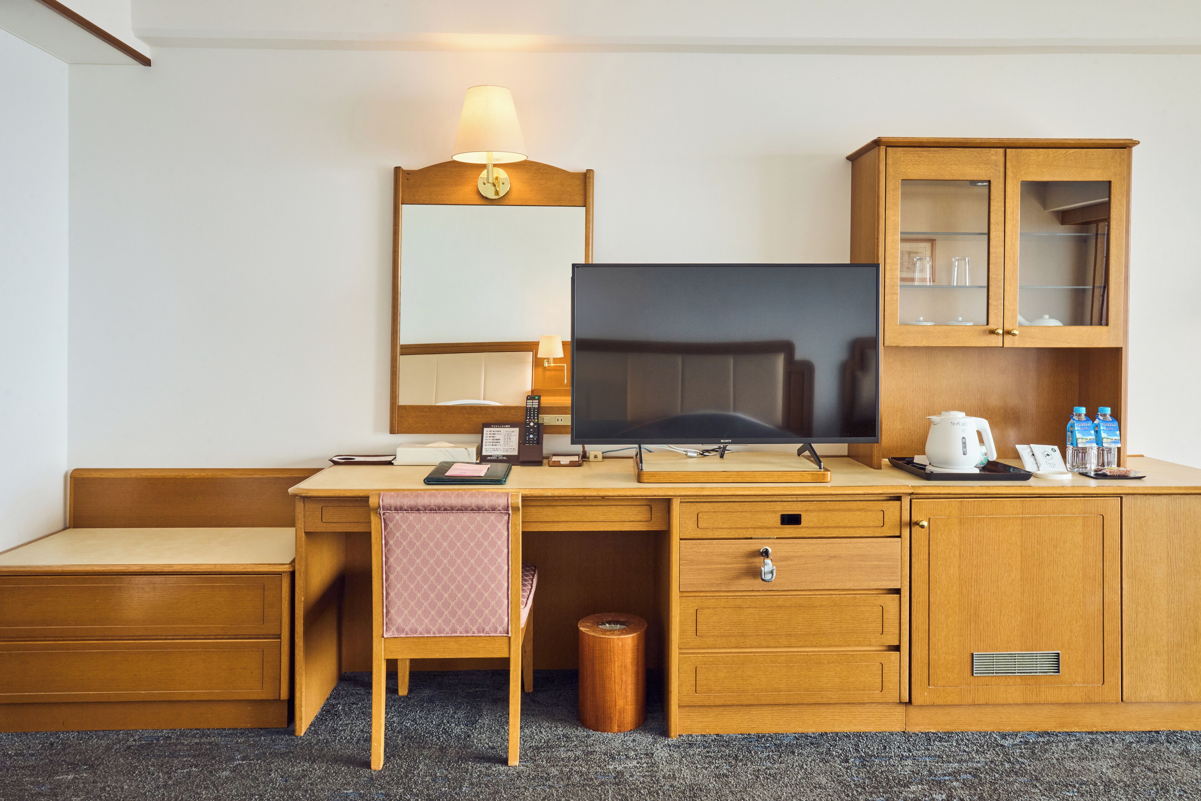 A hotel room with a wooden desk, a television, a cabinet with glass doors, and a small trash can.