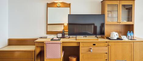 A hotel room with a wooden desk, a television, a cabinet with glass doors, and a small trash can.