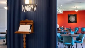 Een receptiegedeelte met een donkerblauwe muur, een houten etagère met een menukaart, en een rode muur waarop een rond kunstwerk hangt.