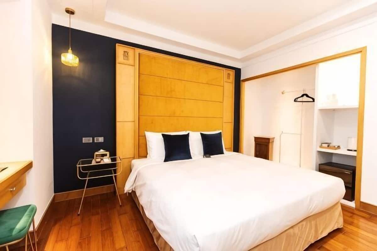 A hotel room with a wooden headboard, a bed with white bedding, a small table with a chair, and a wardrobe.