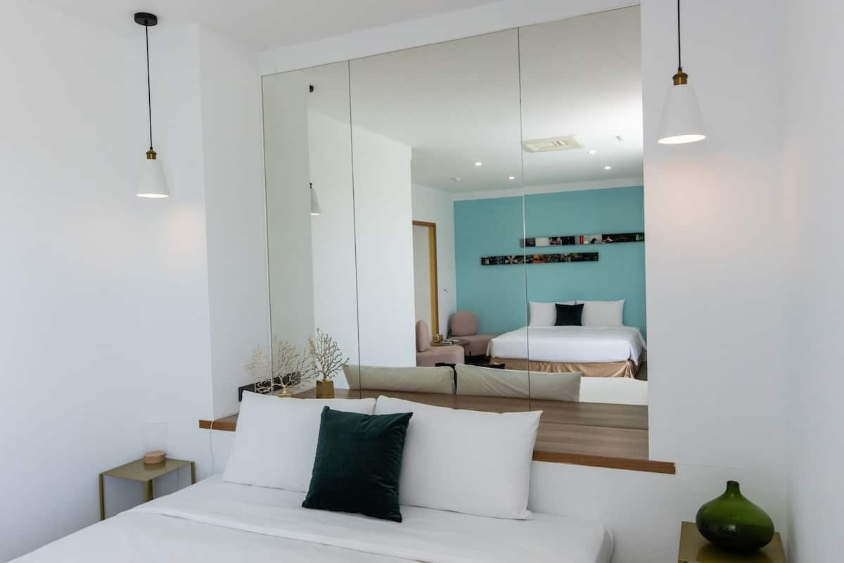 A modern hotel room with a large mirror, a bed with white linens, a bedside table, and a green vase.