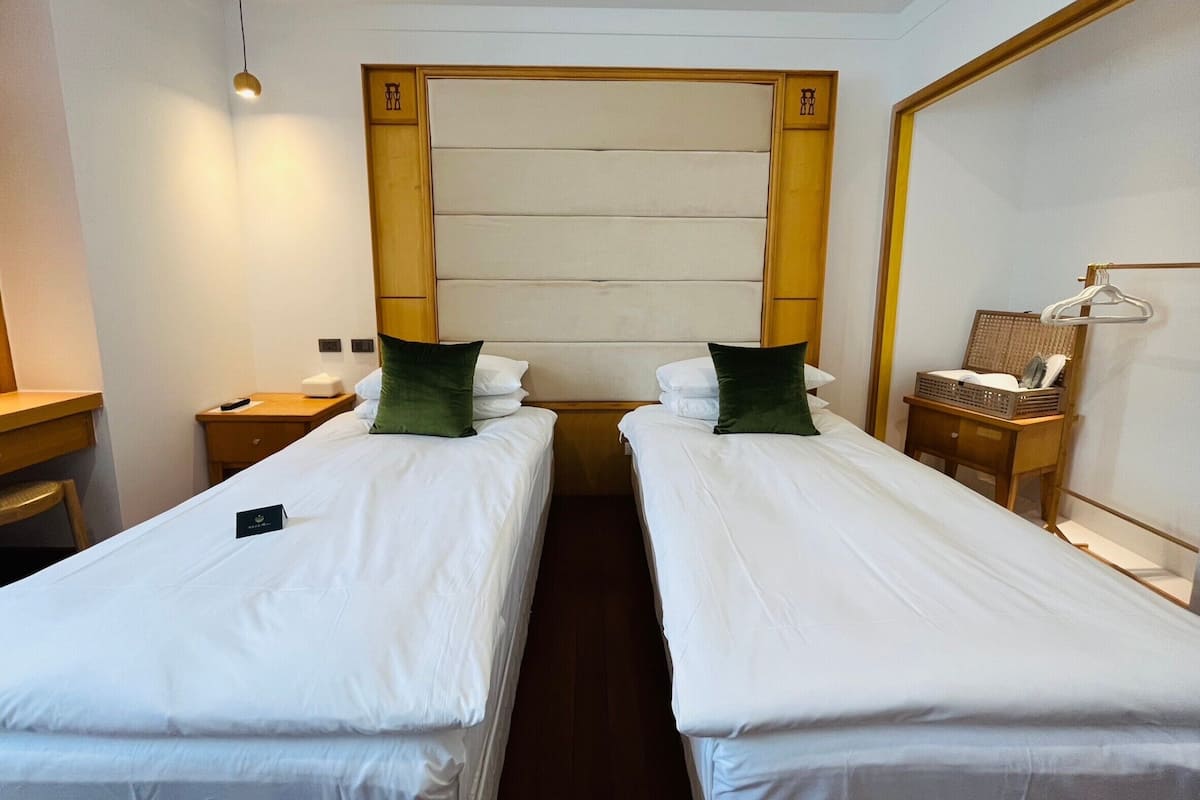 A hotel room with two single beds, a wooden headboard, a bedside table with a lamp, and a hanging rack with clothes.
