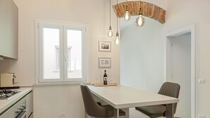 A modern kitchen with a dining area, featuring a white table, two chairs, a hanging light fixture, and a window with blinds. - Homiday- House L -Wood beams and comfort by the sea (Livorno)
