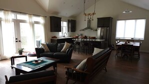 Amplia sala de estar con sofá, mesa de centro y mesa de comedor. Cuenta con una zona de cocina con estufa, fregadero y refrigerador. La habitación tiene ventanas grandes y una lámpara de techo.