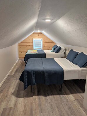 A small, attic-style bedroom with two beds, a wooden sloped ceiling, and a window with blinds.