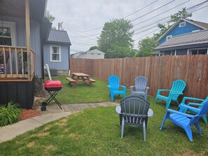 A backyard with a wooden deck, a barbecue grill, a picnic table, and several Adirondack chairs.