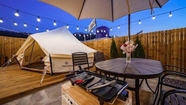 Un espace extérieur aménagé avec une tente, une table et des chaises, des guirlandes lumineuses et une enseigne indiquant « HÔTEL ».