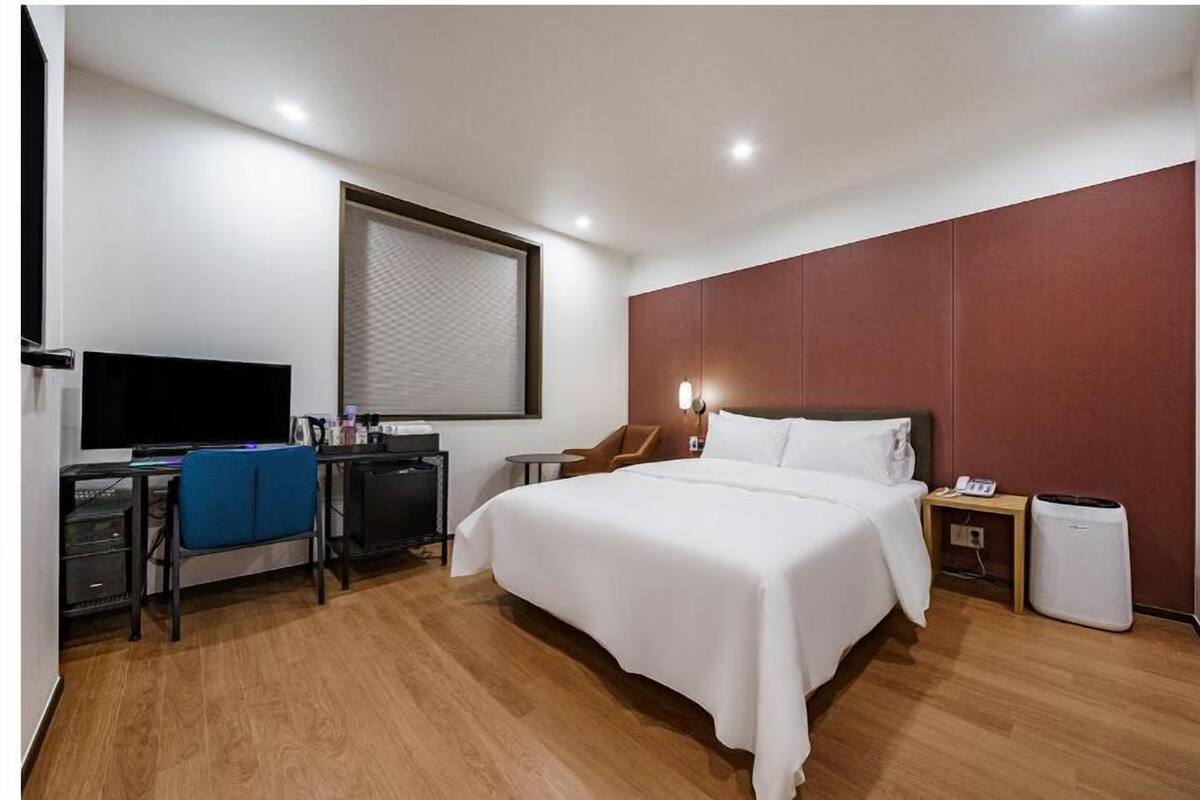 Habitació d'hotel moderna amb un llit gran, una taula de treball amb un ordinador, una televisió i una finestra amb persianes.