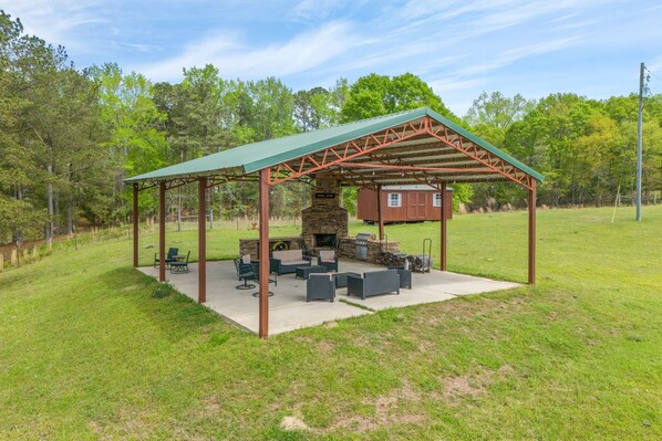 A covered outdoor seating area with a stone fireplace, surrounded by a metal frame and a green roof.