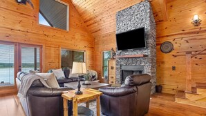 A cozy cabin interior with a stone fireplace, leather sofas, and a television mounted on the wall.