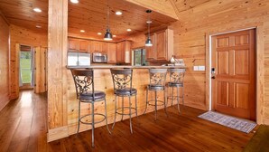 A wooden interior with a kitchen area, bar stools, and a door.