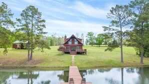 A cabin with a dock extending into a calm lake, surrounded by trees and a grassy field.