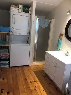Un espace buanderie équipé d’un lave-linge et d’un sèche-linge, un plancher en bois, une salle de bain avec un lavabo et un miroir, ainsi qu’un porte-serviettes.