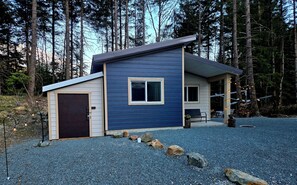 A modern two-story cabin with a dark blue exterior, a front porch, and a gravel driveway.