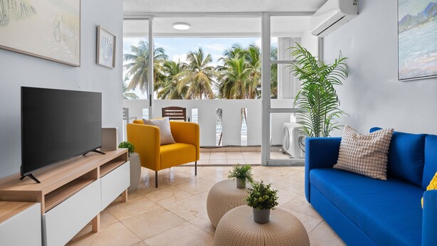 A modern living room with a TV, yellow and blue sofas, and a balcony with palm trees.