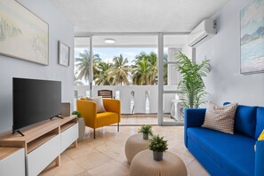 Una sala de estar moderna con televisor, sofás amarillos y azules, y un balcón con palmeras.