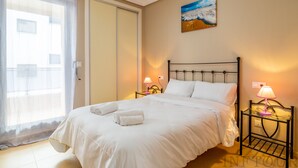 Un lit bien fait, avec du linge de lit blanc et deux serviettes enroulées, une table de chevet avec une lampe et un tableau accroché au mur.