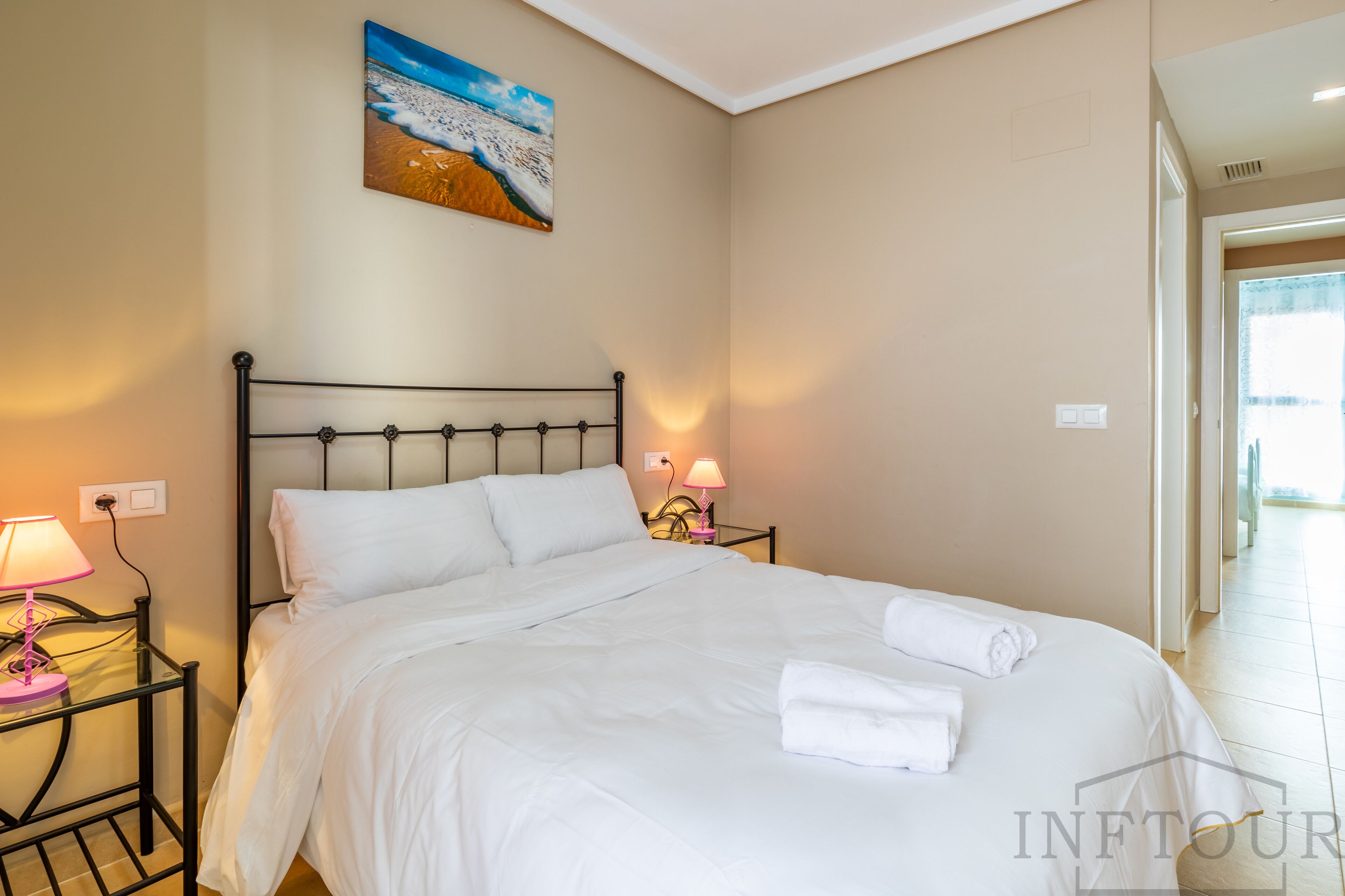 Un lit bien fait, avec du linge de lit blanc et des oreillers, une lampe de chevet et un tableau représentant une scène de plage accroché au mur.