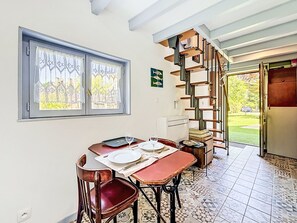 A dining area with a table set for two, a window with lace curtains, a staircase, and a view of a garden.