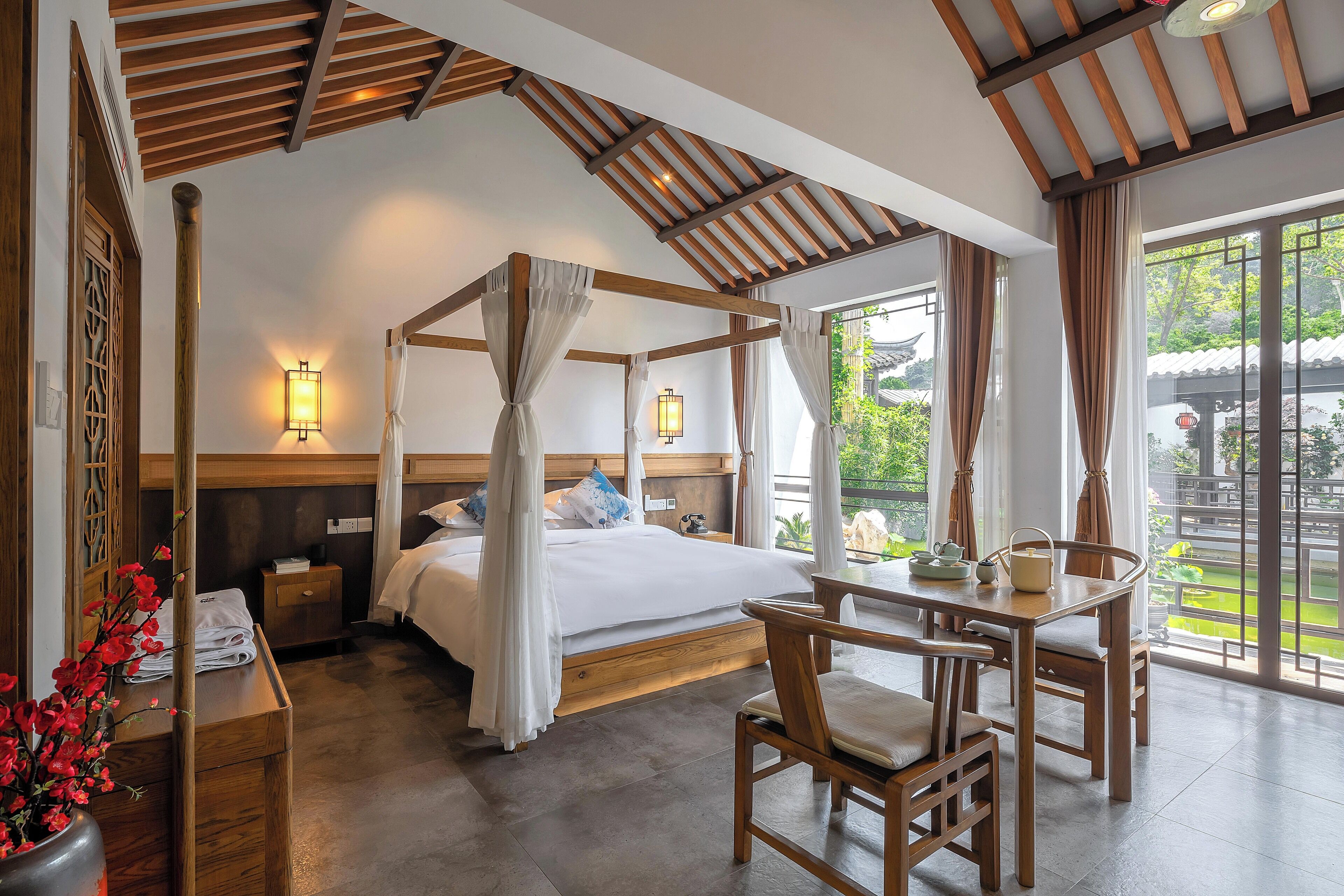 A four-poster bed with white linens, a wooden nightstand, a table with chairs, and a view of greenery outside.