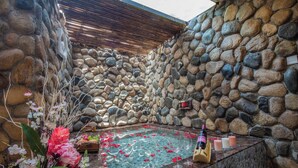 Zona de spa con paredes de piedra, una bañera de hidromasaje, flores y una botella de vino.