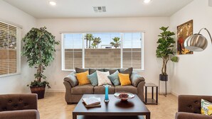 Un salon comprenant un canapé, une table basse et deux fauteuils. On y trouve une grande fenêtre avec des stores, une plante en pot et un tableau accroché au mur.