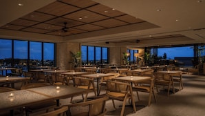 A spacious indoor dining area with multiple tables and chairs, large windows offering a view of the cityscape at dusk, and a ceiling with recessed lighting. - THE NEST NAHA (Naha)
