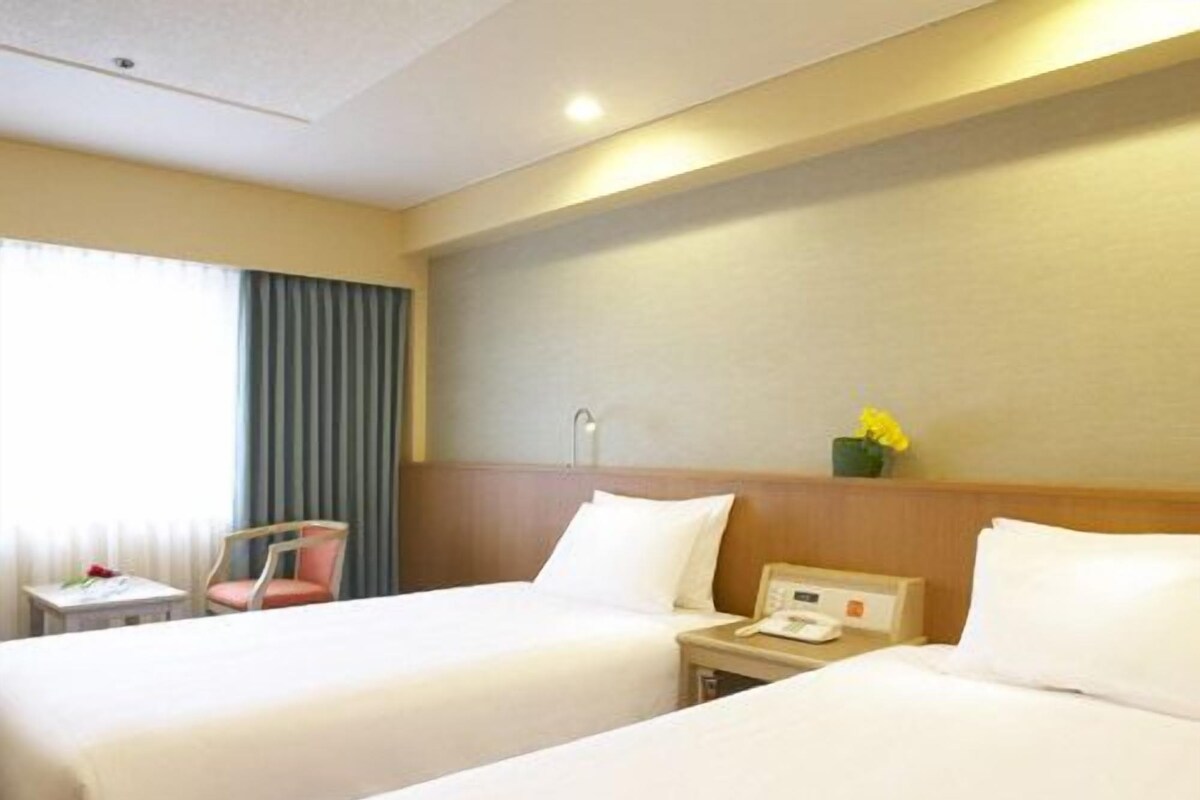 A hotel room with two beds, a bedside table with a lamp, a chair, and a window with curtains.