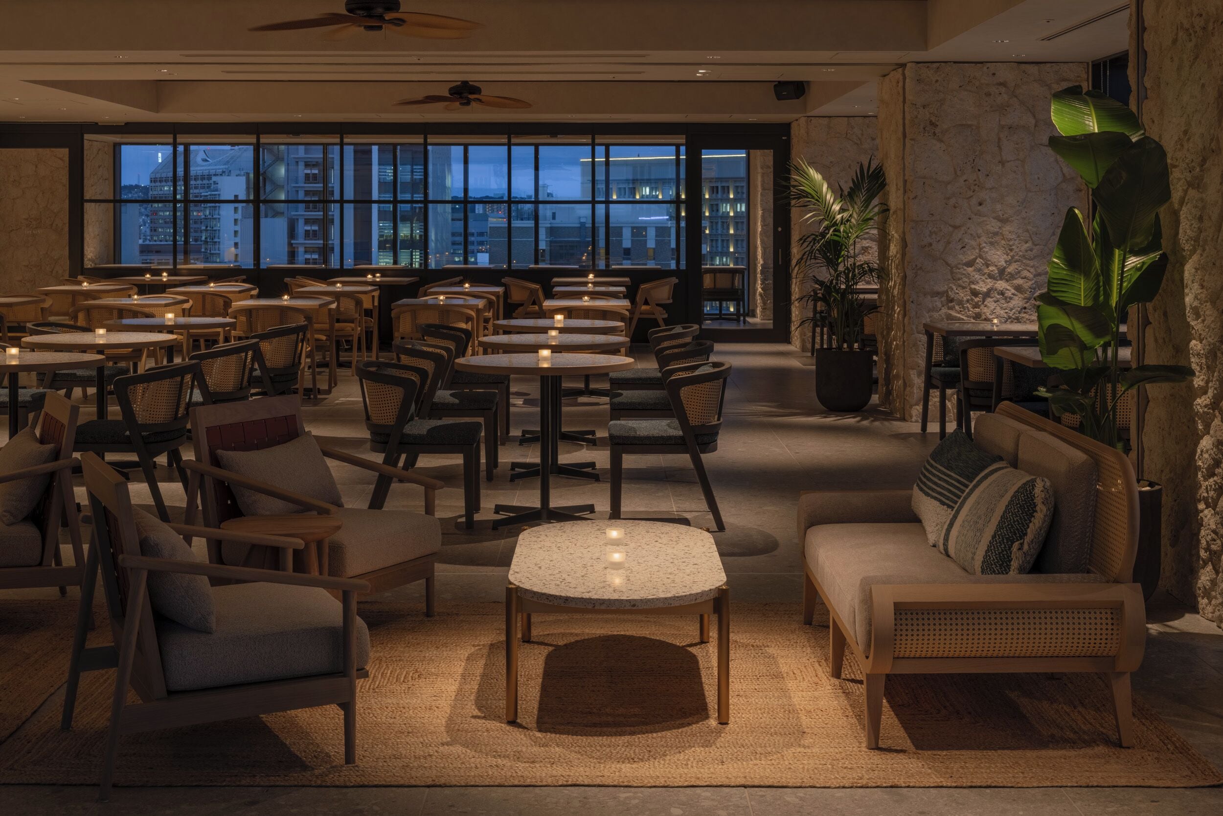 A modern dining area with round tables and chairs, a sofa, and large windows offering a city view.