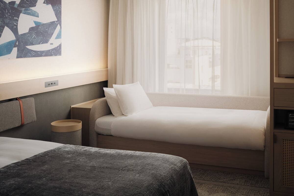 A hotel room with a bed, a bedside table, a lamp, a window with sheer curtains, and a wall-mounted artwork.