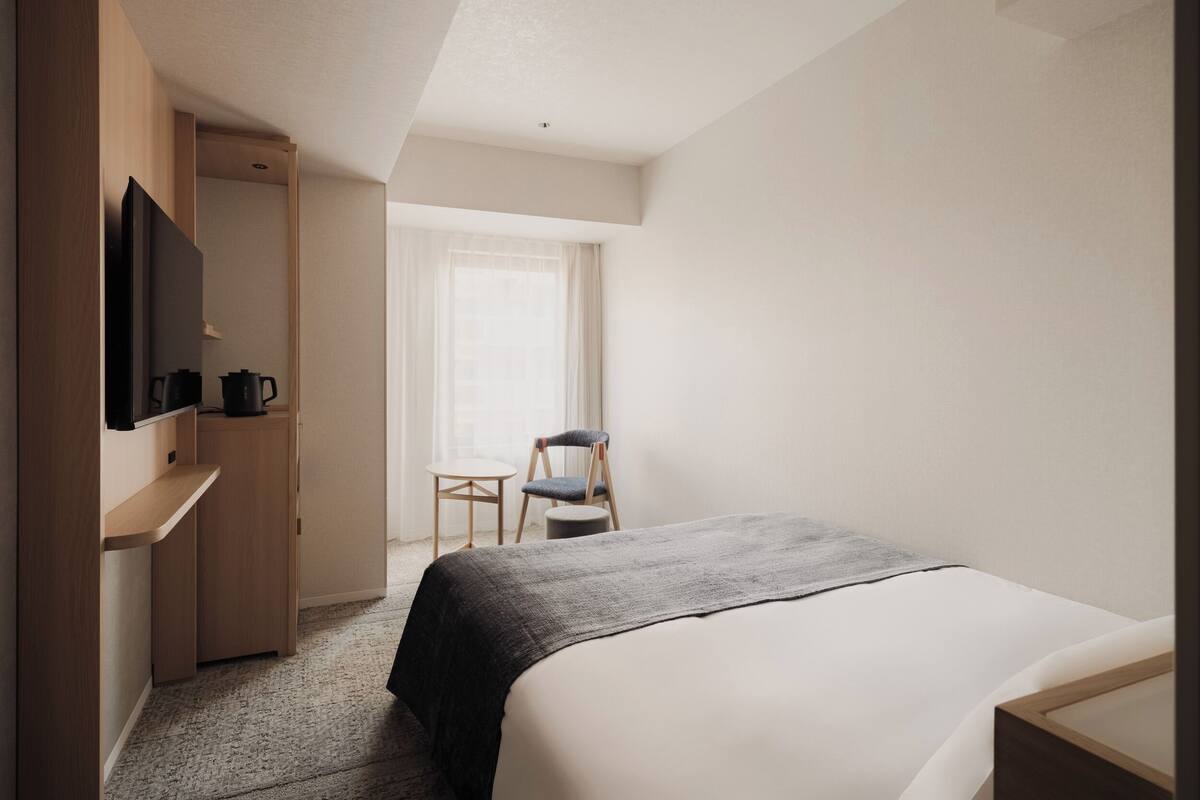 A hotel room with a bed, a television on a wooden stand, a small table with a chair, and a window with curtains.