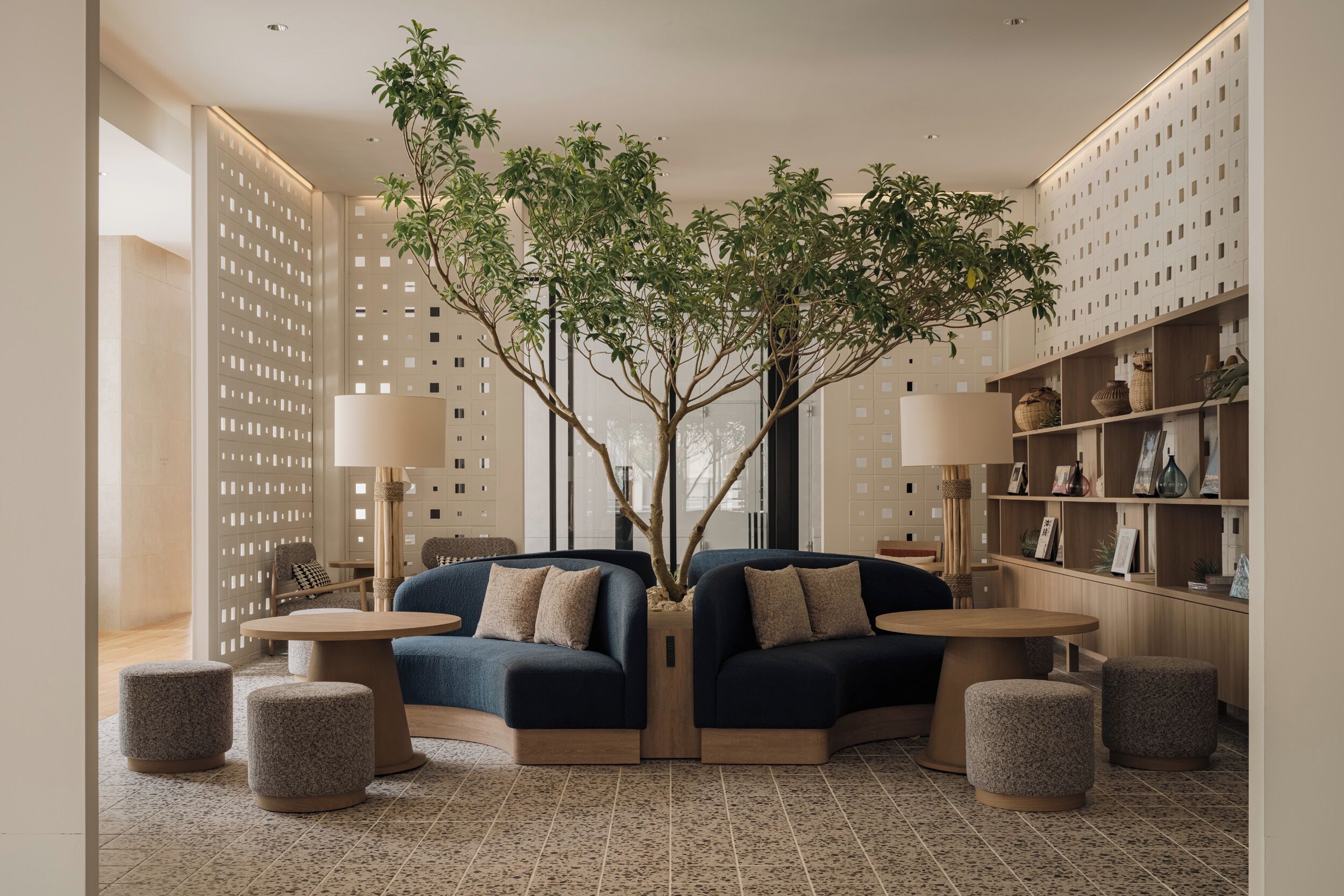 A modern lounge area with two dark blue sofas, round wooden tables, and a central tree planter.