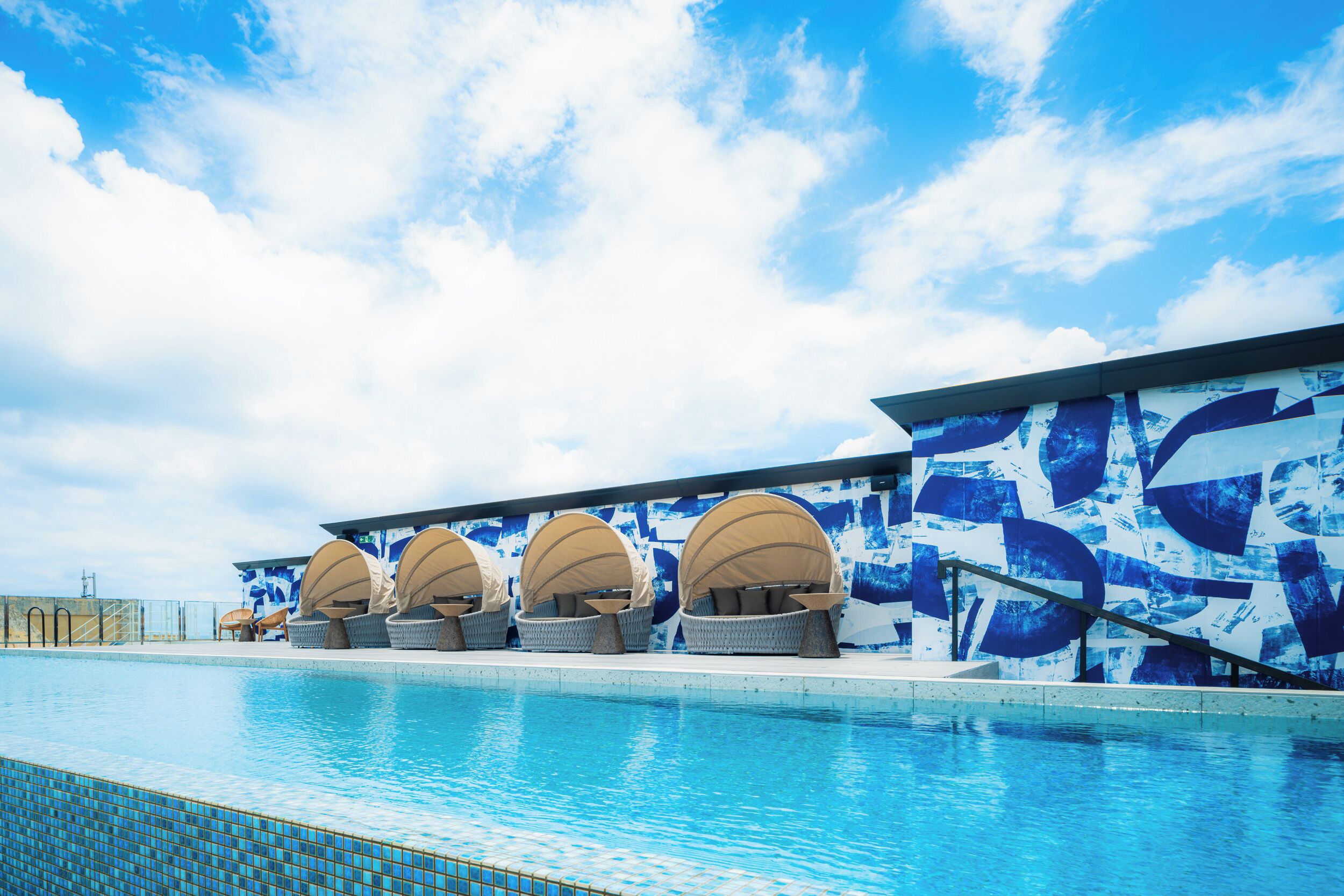 Una piscina con sillas de descanso y sombrillas, un suelo de baldosas y una pared con diseños abstractos en azul y blanco.