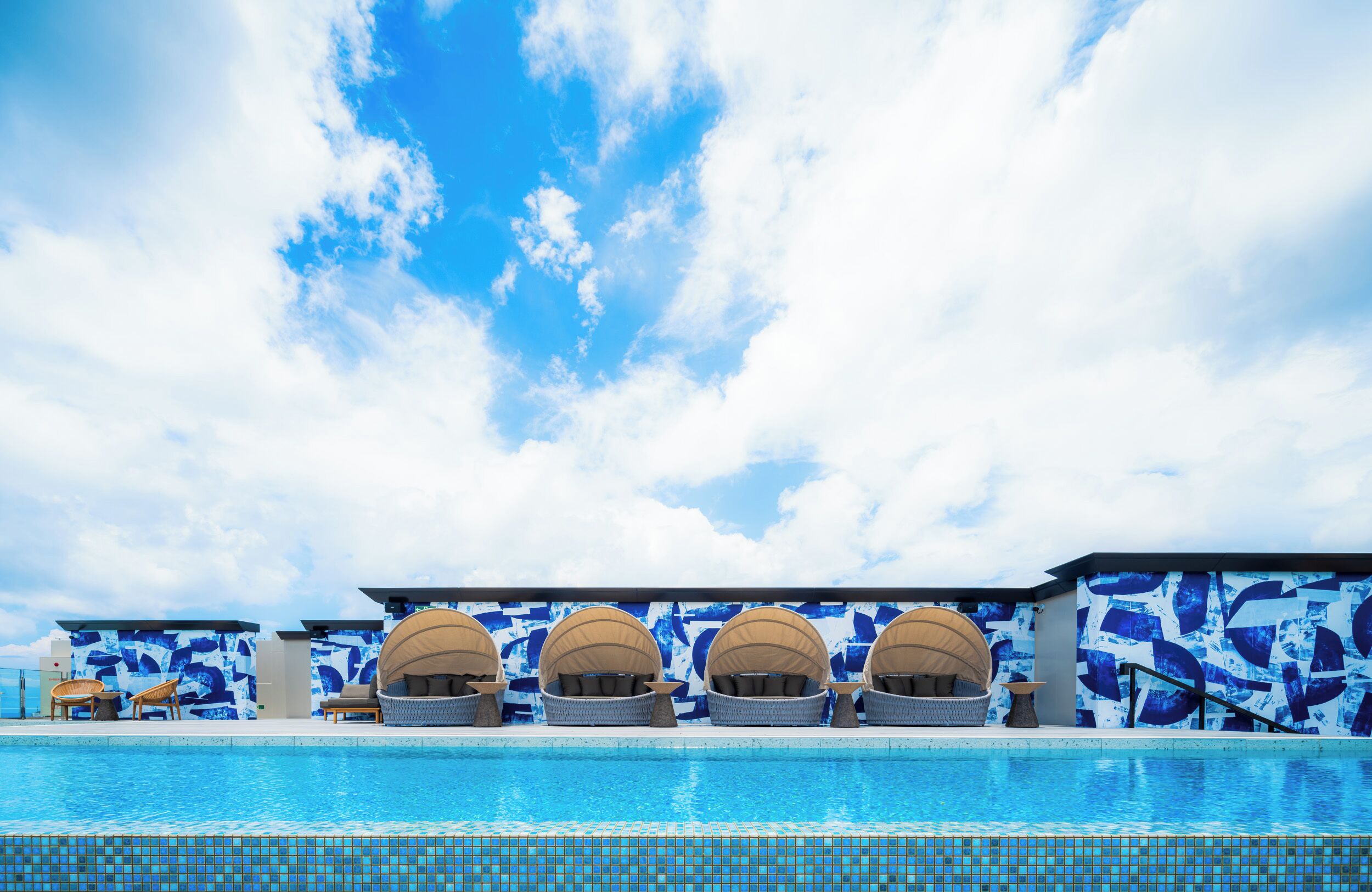A swimming pool with lounge chairs and umbrellas, a blue and white patterned wall, and a clear sky.