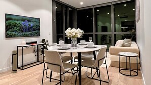 Un comedor moderno con una mesa redonda, cuatro sillas y un jarrón con flores blancas. Hay un televisor de pantalla plana montado en la pared, un ventilador de techo y ventanas grandes que aportan luz natural.