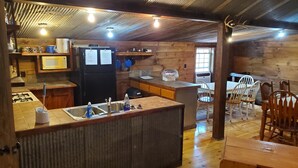 Uma cozinha rústica com paredes de madeira, uma geladeira preta, micro-ondas e pia. Há um espaço de refeições com cadeiras e mesa de madeira.