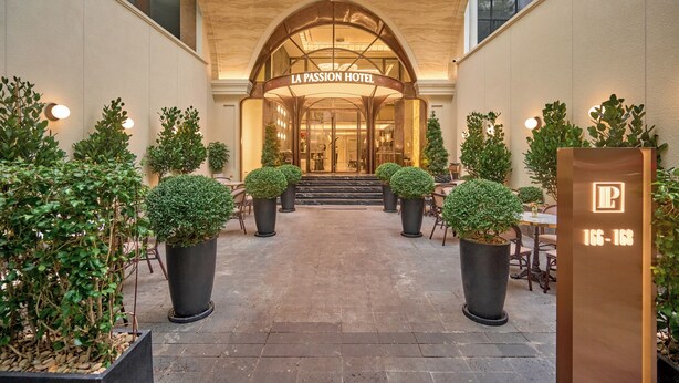 The entrance of La Passion Hotel with a grand arched doorway, potted topiary plants, and a sign displaying the hotel's name and address.