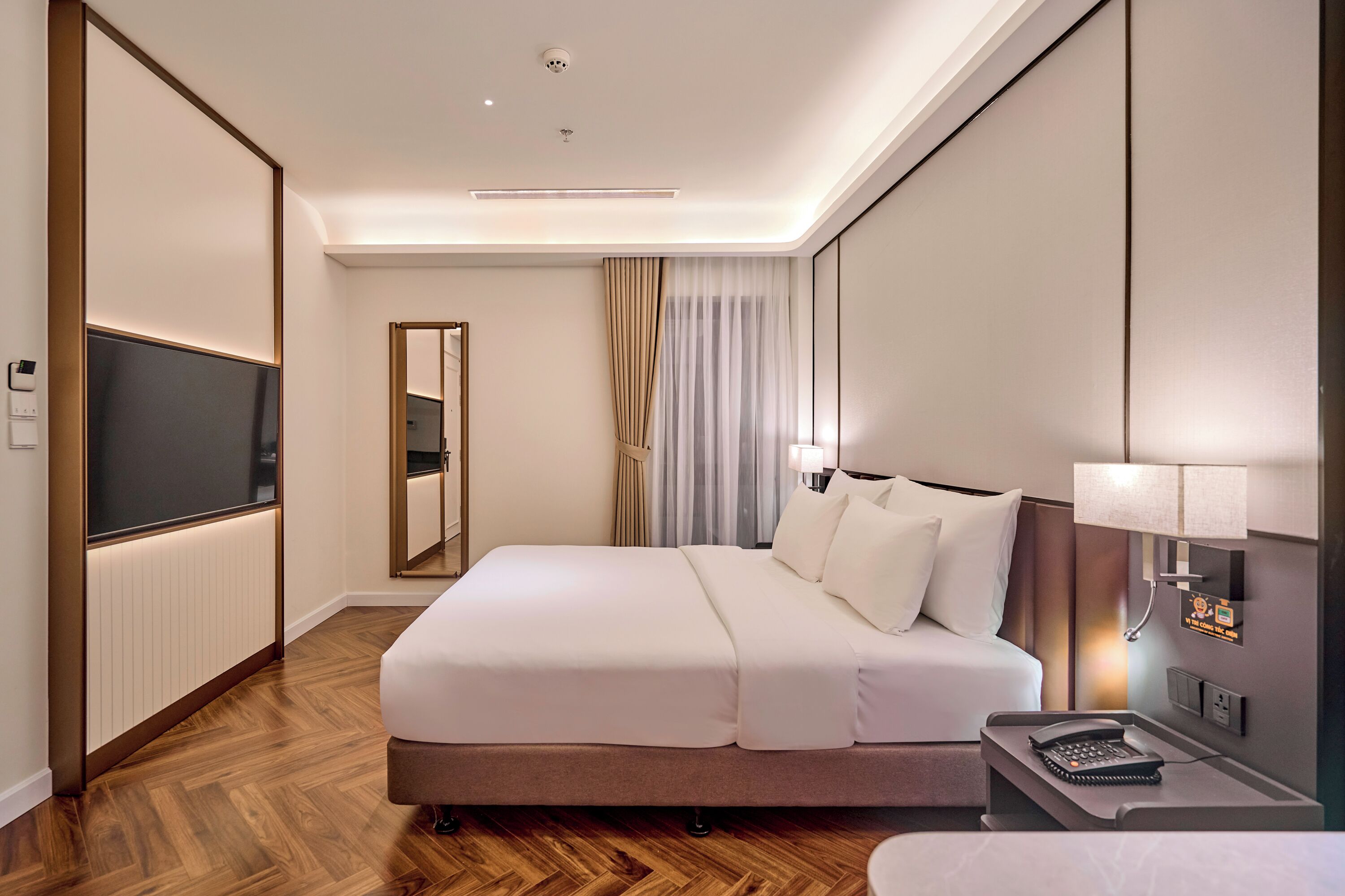 A modern hotel room with a large bed, a flat-screen TV, a bedside table with a lamp, and a mirror.