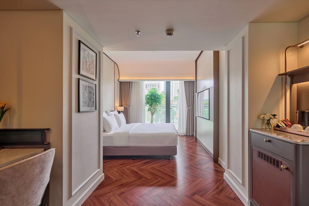 A hotel room with a bed, a desk, a dresser, and a large window with a view of buildings and trees.